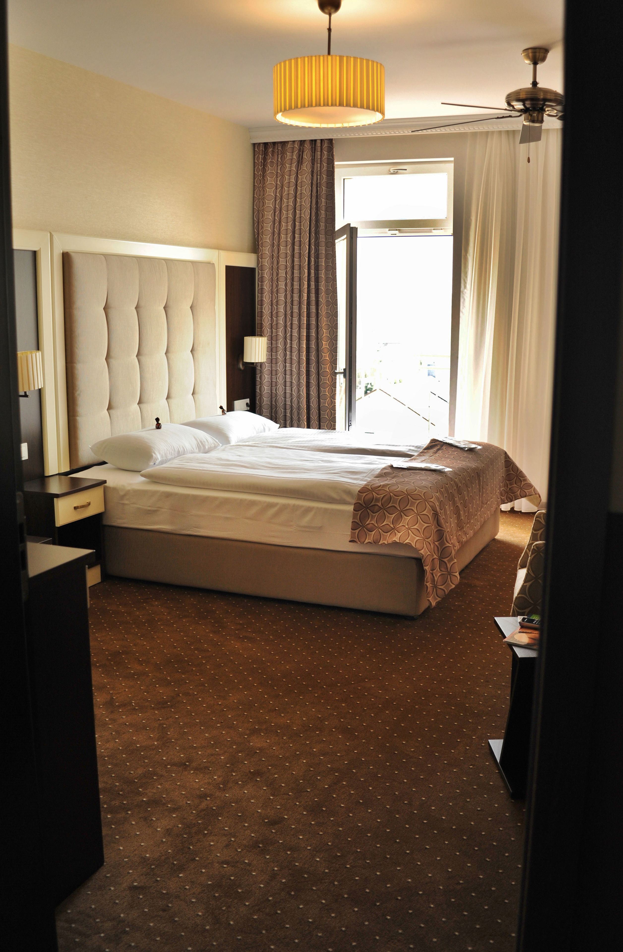 A hotel room with a large bed, a ceiling fan, a window with curtains, and a nightstand with a lamp.