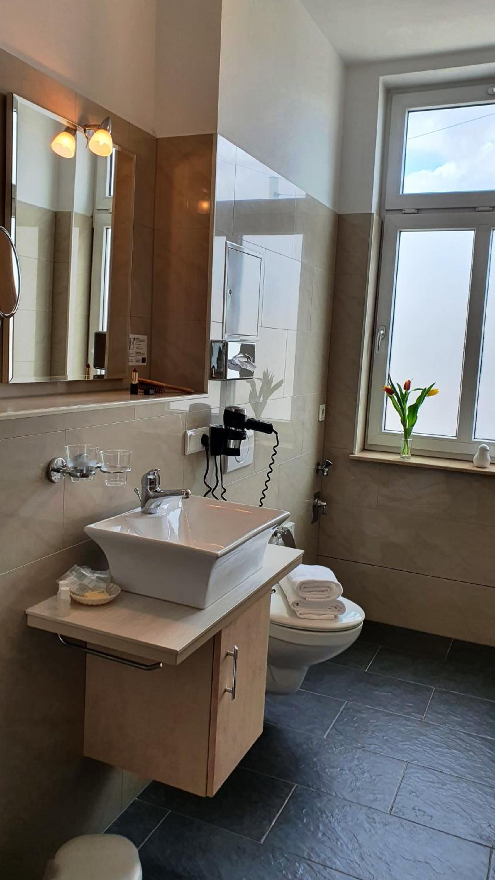 Une salle de bain moderne avec un grand miroir, un lavabo blanc, des toilettes et une fenêtre donnant sur le ciel.