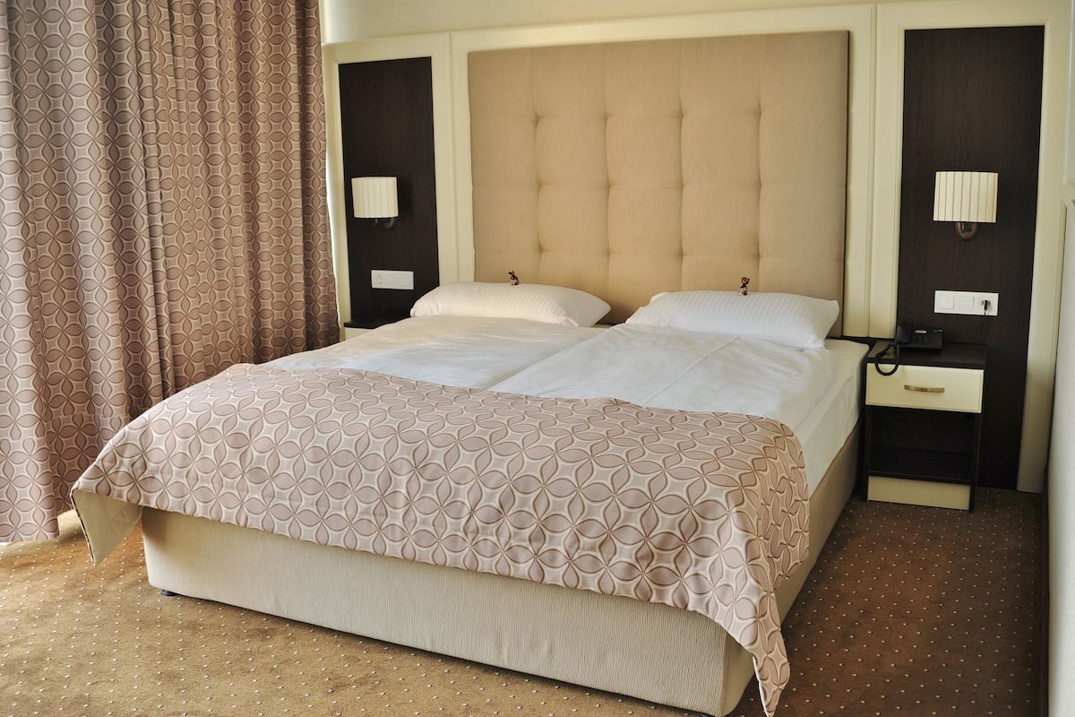 A double bed with a patterned bedspread, two bedside tables with lamps, and a curtain on the left.