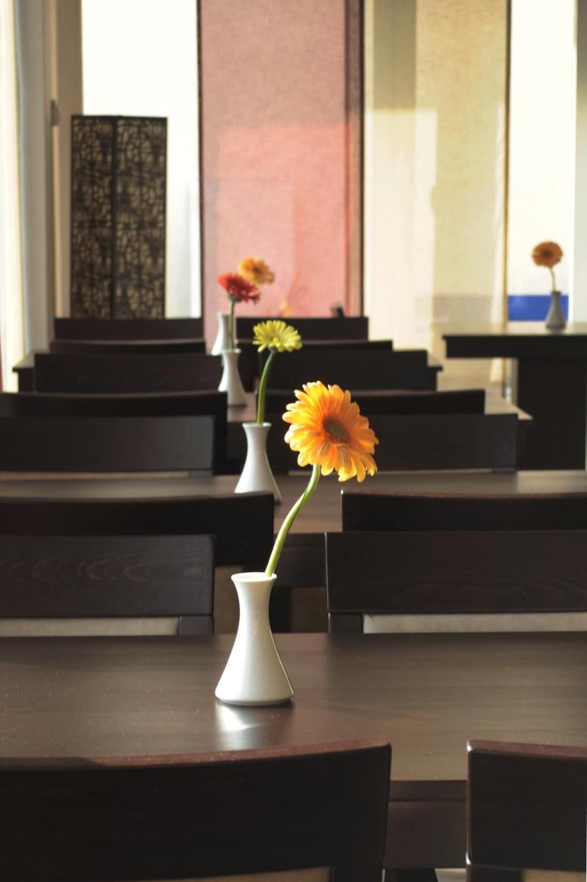 A dining area with tables and chairs, vases with flowers, and a large window in the background.