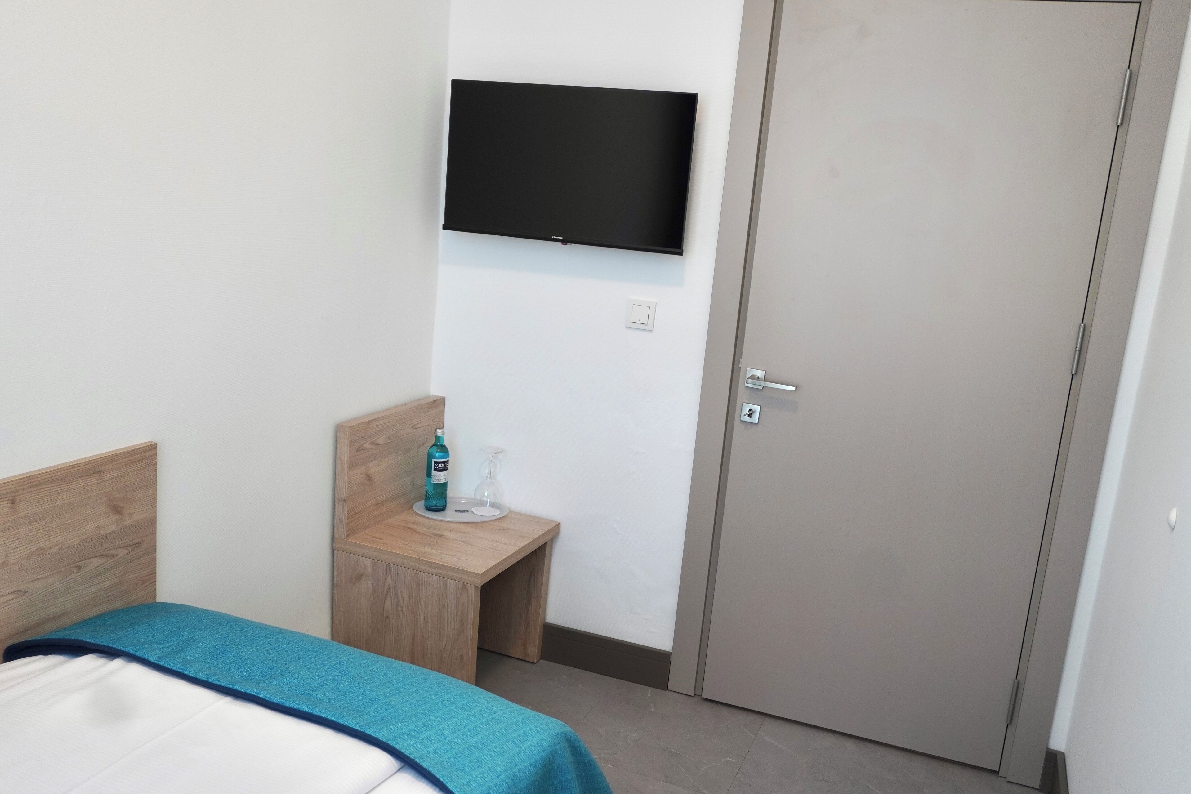A hotel room with a bed, a small wooden nightstand, a television mounted on the wall, and a door.