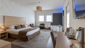 Una habitación de hotel moderna con una cama grande, un escritorio, una silla, una mesita, un sofá, un televisor y una ventana con persianas.