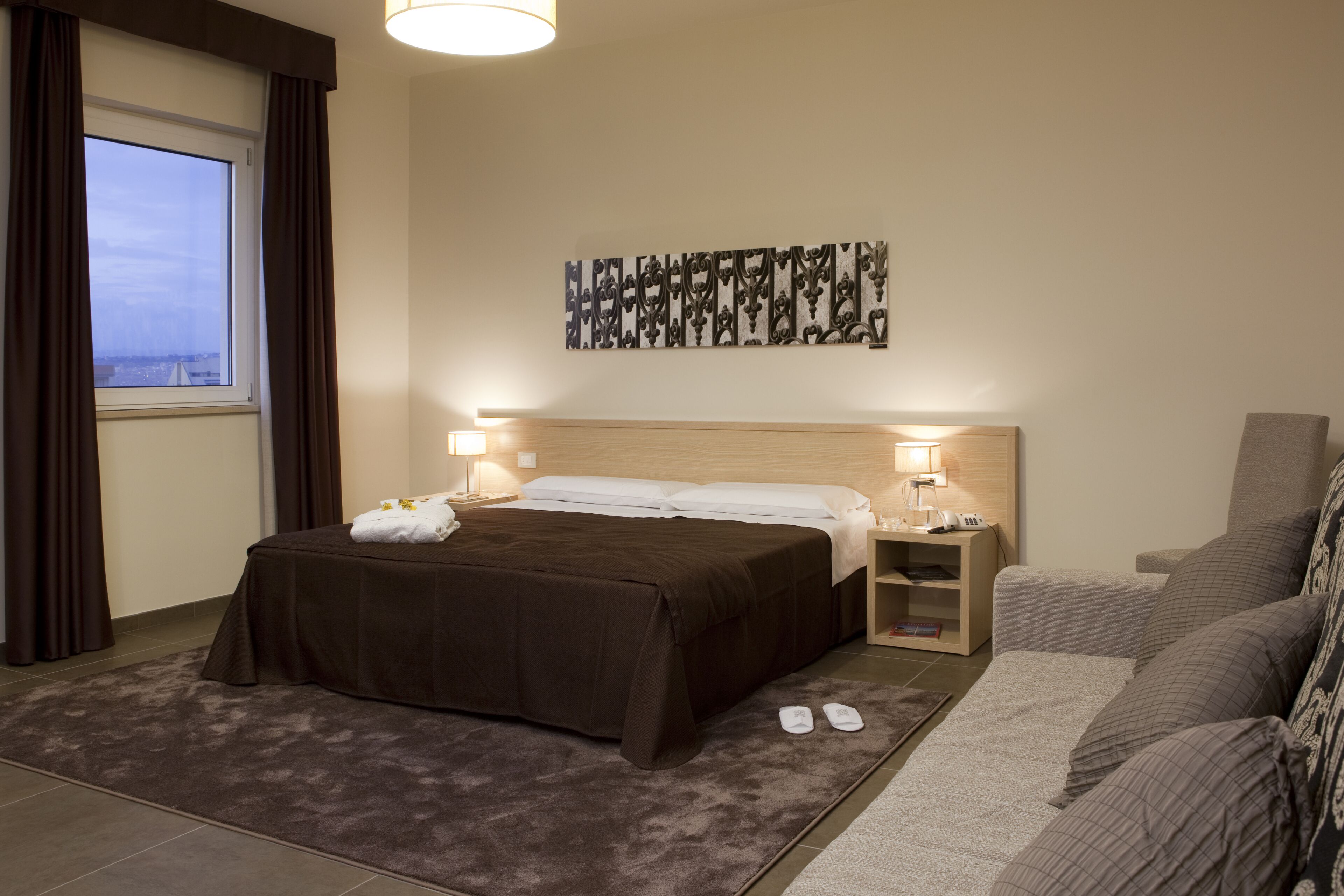 A hotel room with a bed, bedside tables, a sofa, a window with curtains, and a wall art piece.