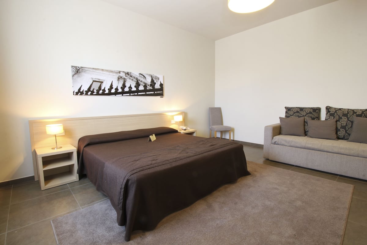 A hotel room with a large bed, a nightstand with a lamp, a sofa with patterned cushions, and a wall-mounted picture.