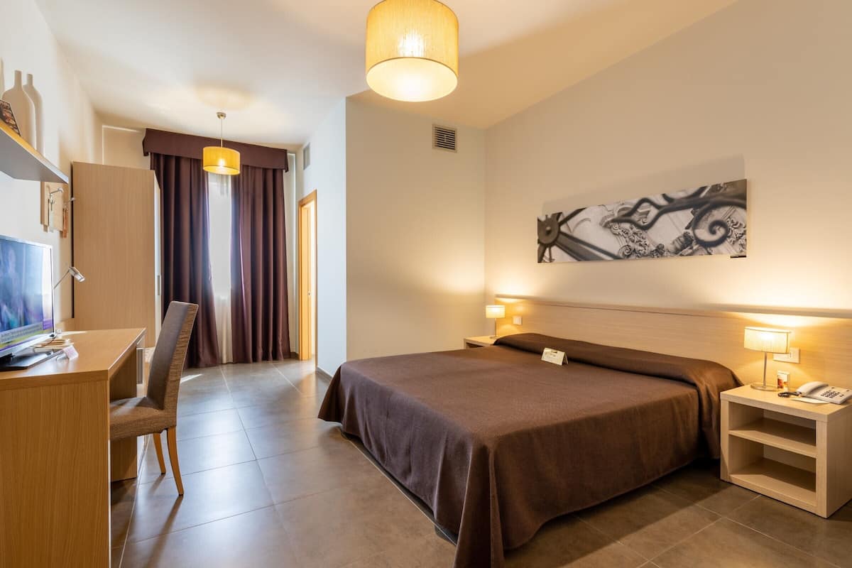 A hotel room with a bed, desk, chair, and a painting on the wall.