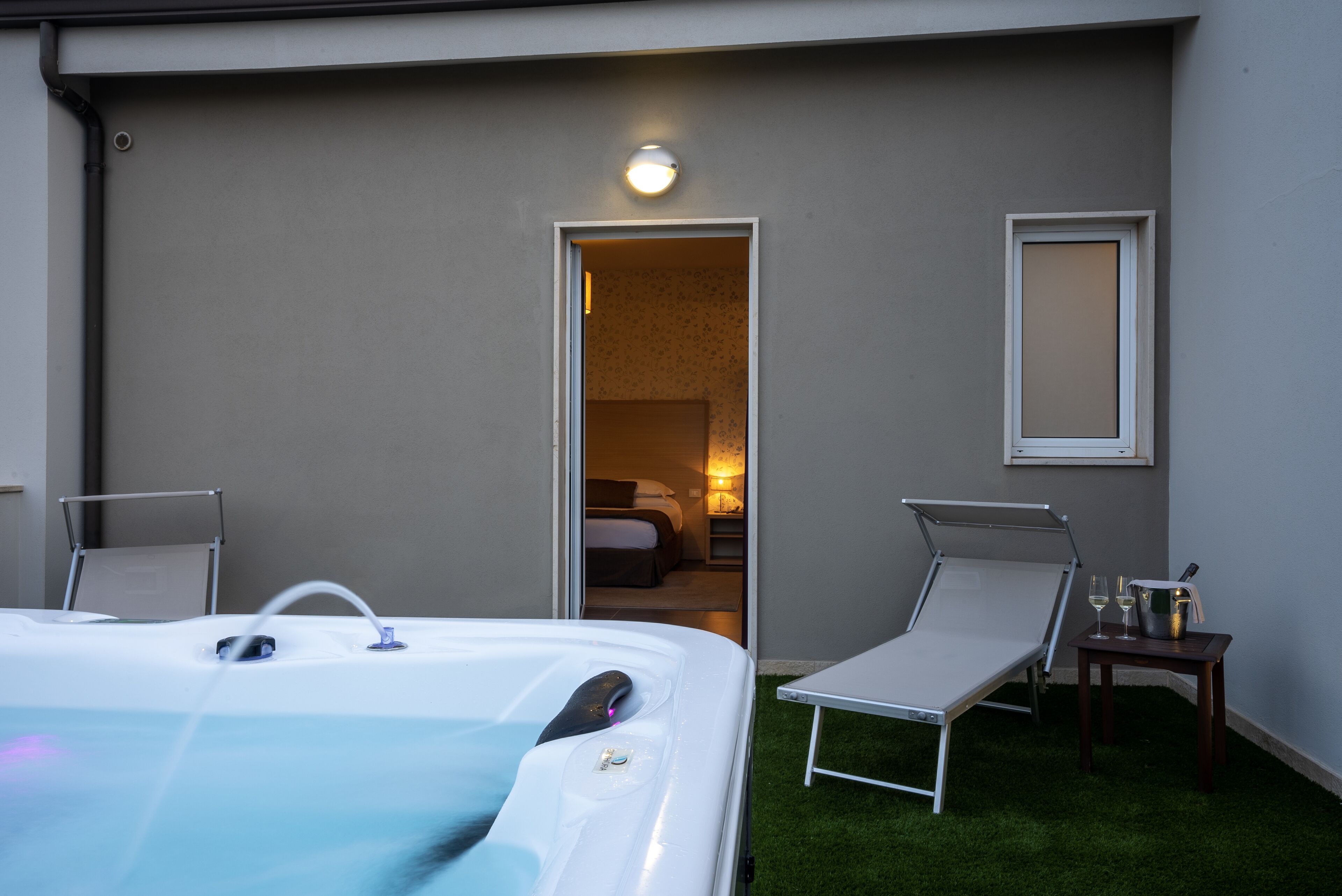 A hot tub with two lounge chairs, a small table with a bottle and glasses, and a door leading to a bedroom.