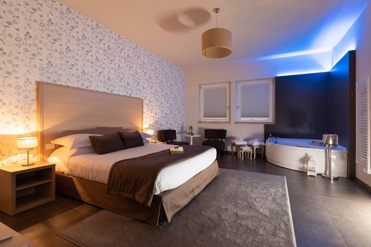 A modern hotel room with a large bed, a desk, a chair, a small table, a sofa, a bathtub, and a wall-mounted TV.