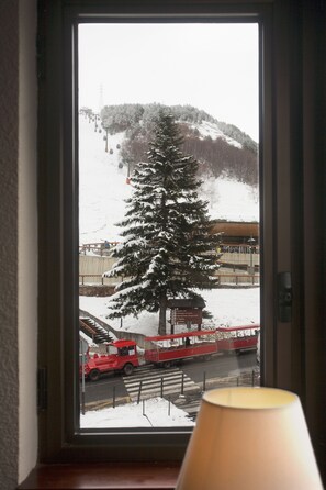 A window view of a snowy mountain landscape with a red vehicle on a road, a large evergreen tree, and a building in the background.