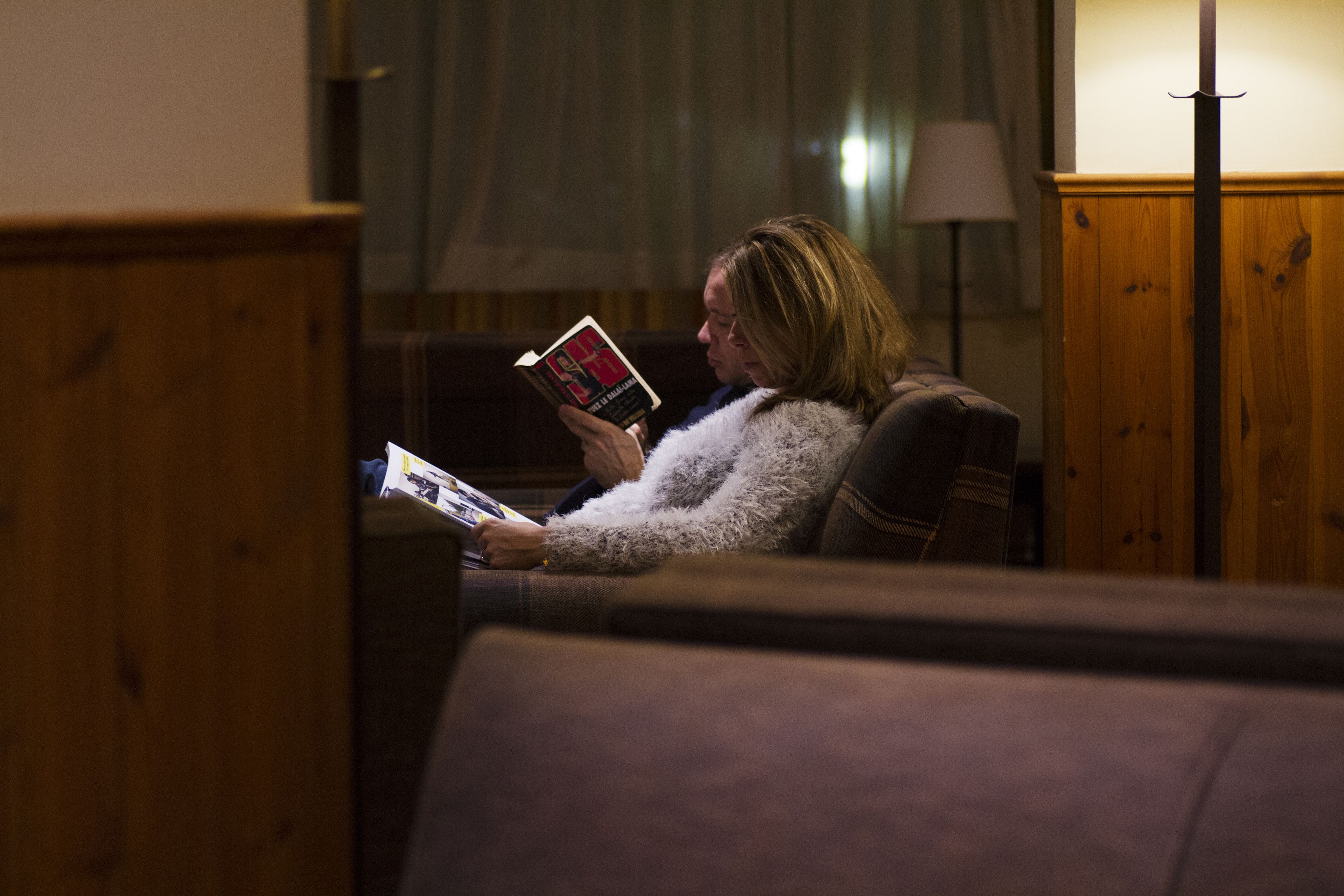 A person sitting on a couch reading a book in a room with wooden furniture and a lamp.