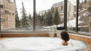 Una persona en un jacuzzi con vistas a árboles nevados y edificios a través de amplias ventanas.