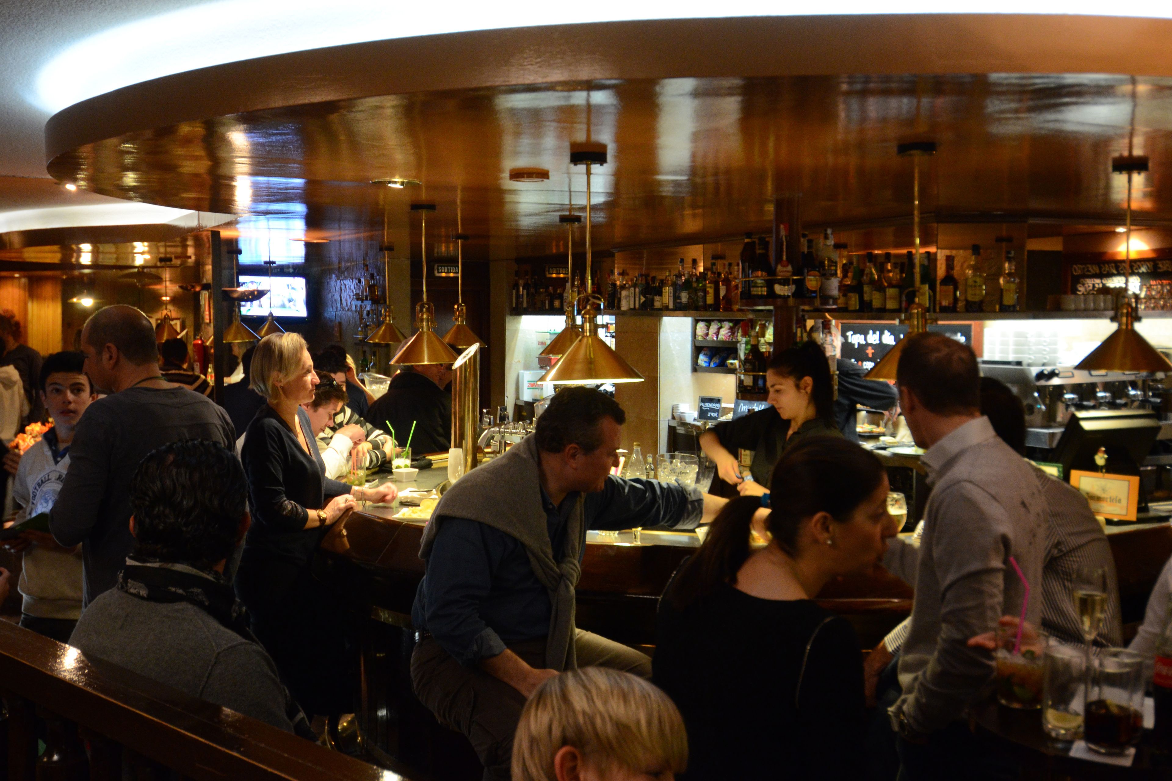 Un bar con clientes sentados y de pie, una barra con un cantinero, y estantes llenos de botellas.
