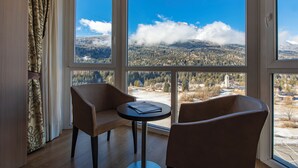 A room with two chairs and a small table, large windows with a mountain view, and patterned curtains.