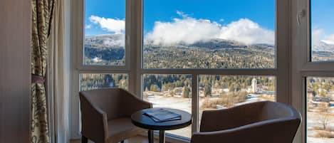 A room with two chairs and a small table, large windows with a mountain view, and patterned curtains.