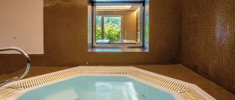 Un bain à remous avec vue sur la verdure, visible à travers une fenêtre, et entouré de murs recouverts de carreaux bruns.