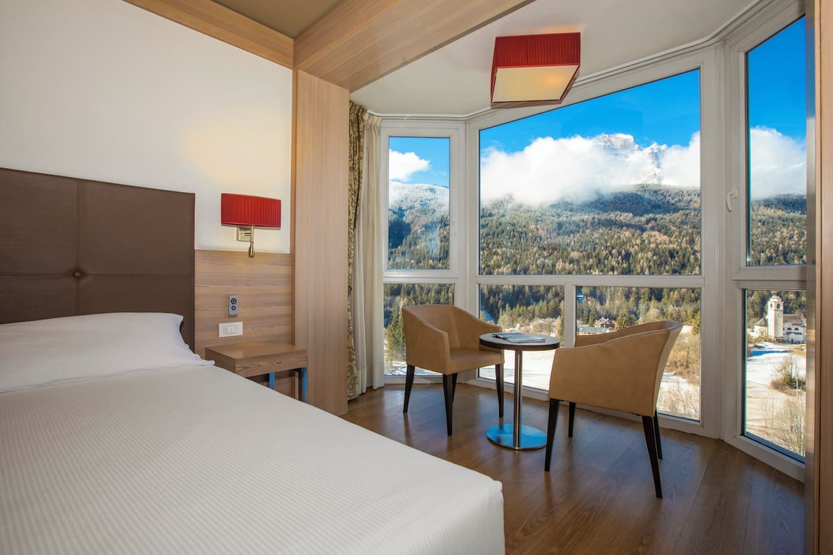 A hotel room with a large bed, two chairs, a small table, and a view of a mountainous landscape through large windows.