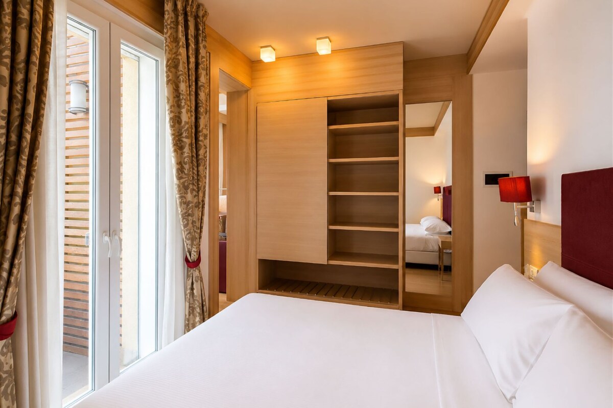 Habitación de hotel con una cama grande, un armario con varios estantes, una mesita de noche con lámpara y una ventana con cortinas.