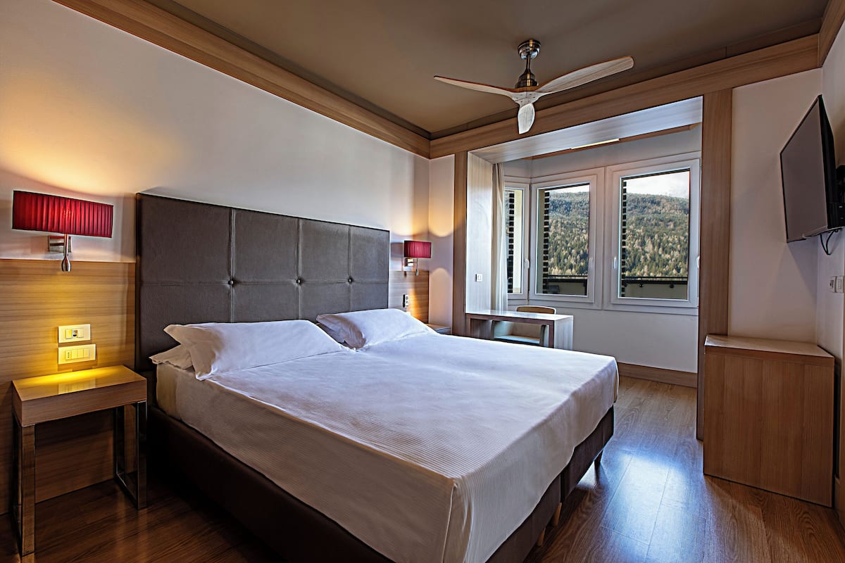 A neatly made bed in a hotel room with a wooden headboard, a nightstand with a lamp, a small table, and a TV on the wall.