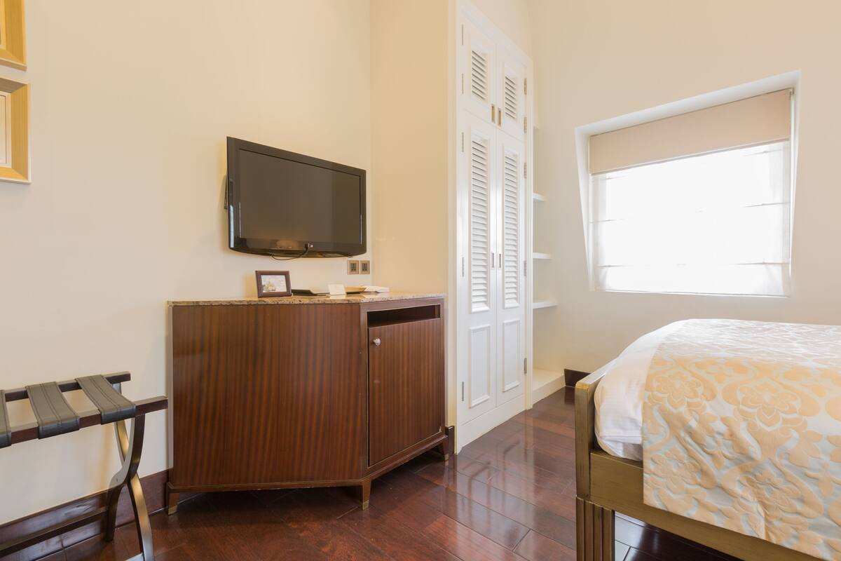 Une chambre à coucher avec un lit, un téléviseur fixé au mur, une armoire en bois et une fenêtre avec des stores.