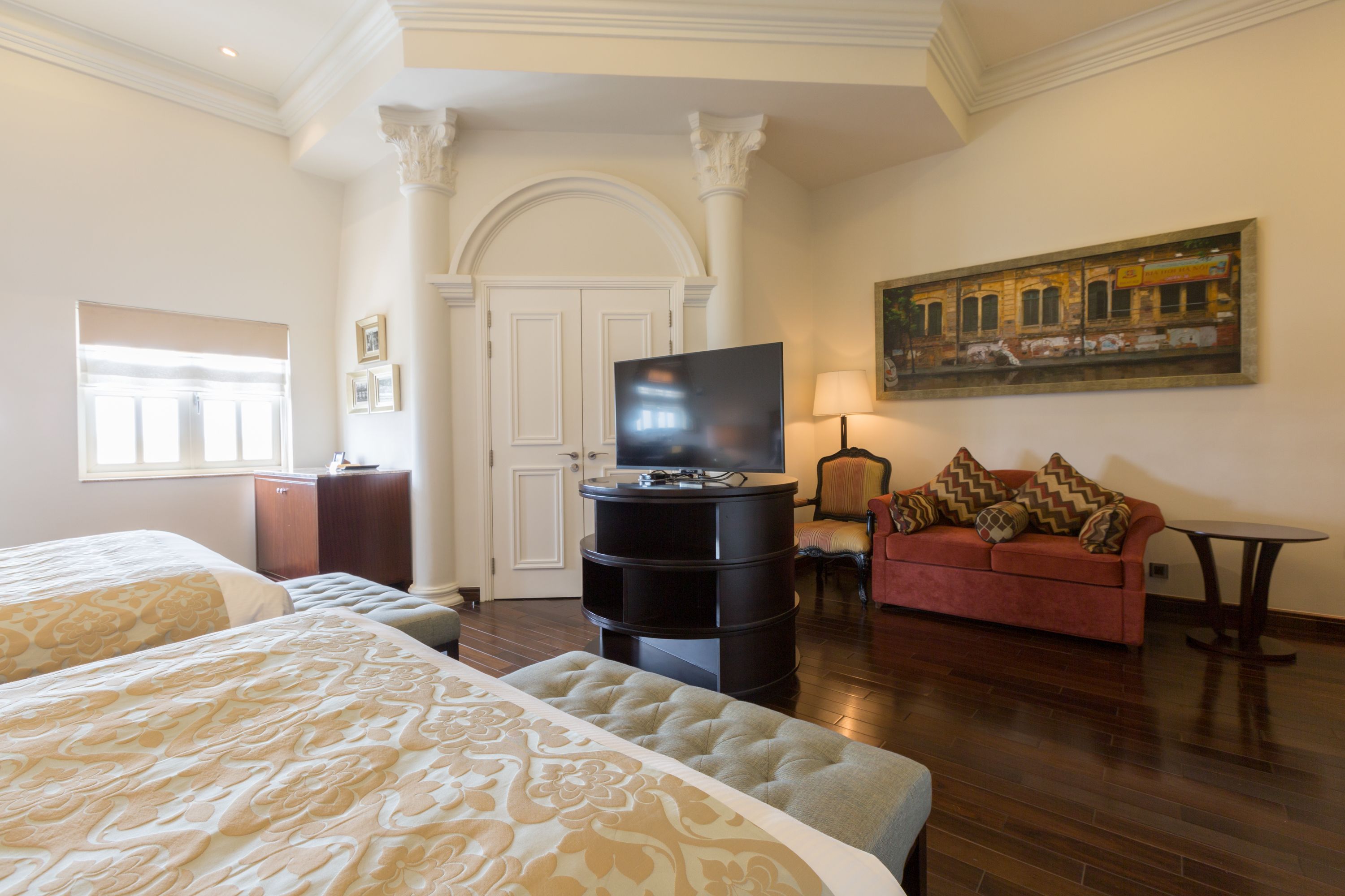 A hotel room with two beds, a TV, a red sofa, and a painting on the wall.