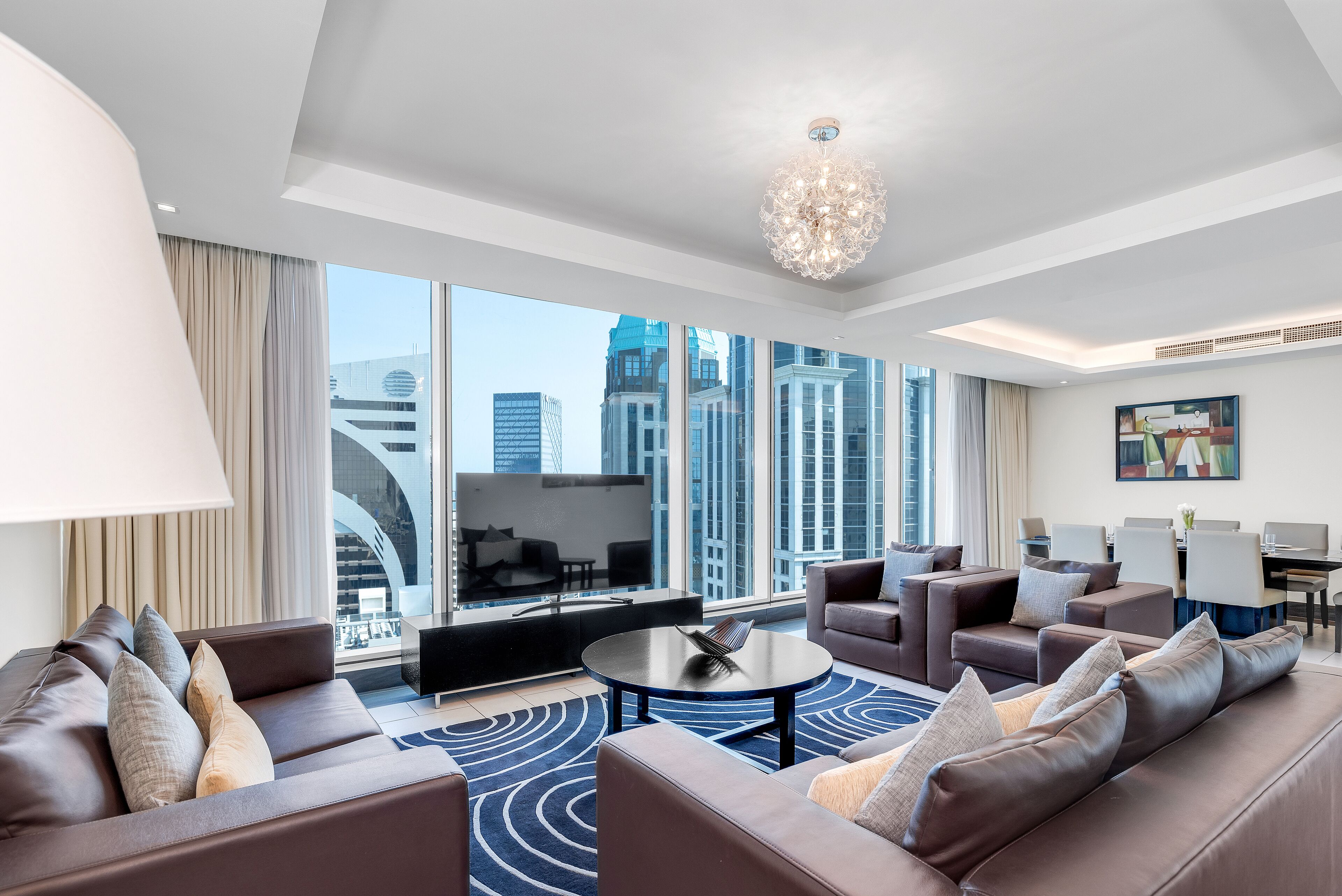 A modern hotel room with a large window offering a city view, a round coffee table, and a seating area with sofas and chairs.