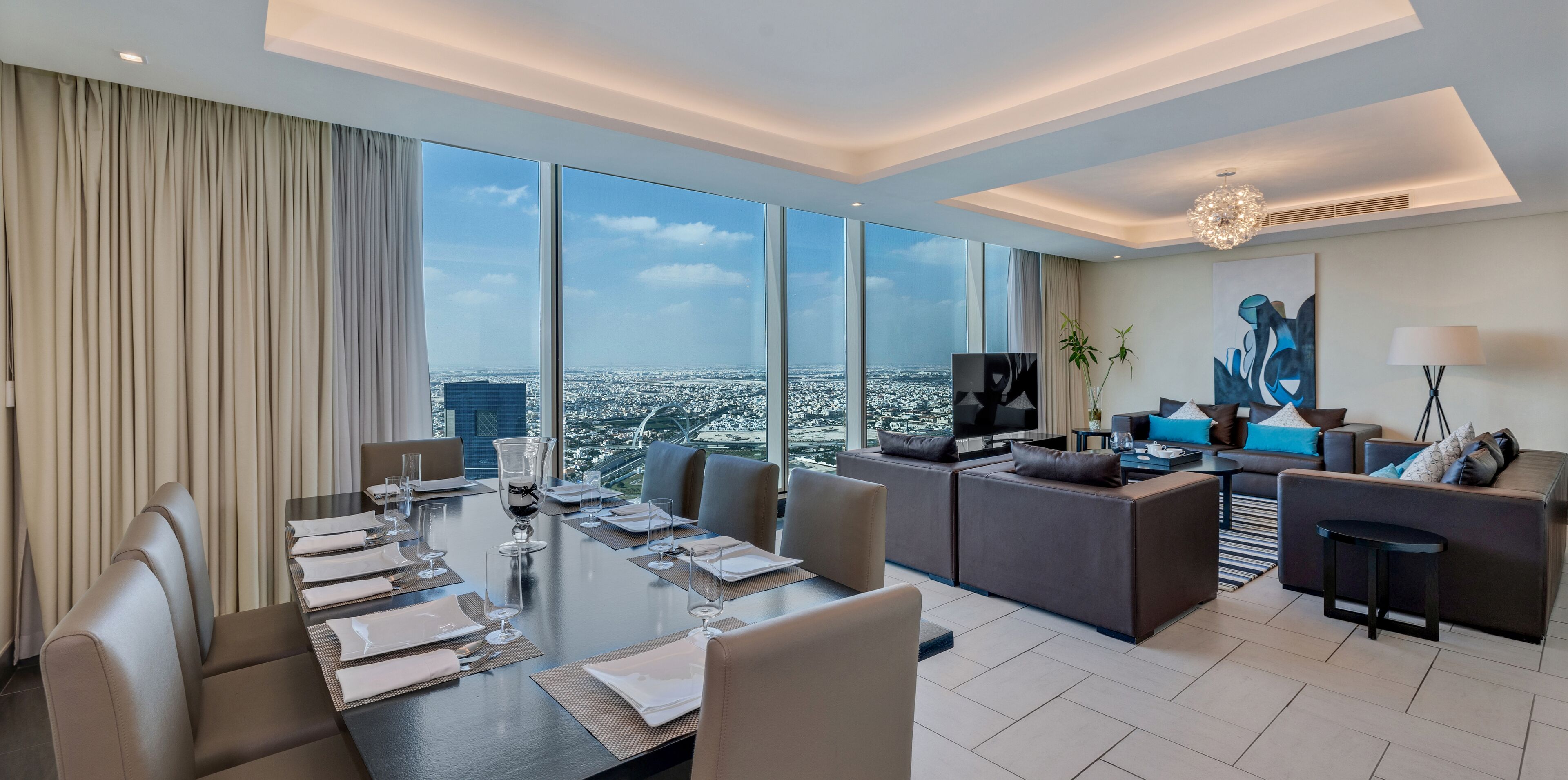 Una habitación de hotel moderna con una gran mesa de comedor preparada para seis personas, una zona de sofás y vistas a la ciudad.