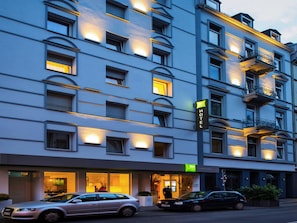 Ein mehrstöckiges Hotel mit beleuchteten Fenstern und Balkonen, einem gut sichtbaren Eingang sowie geparkten Autos.