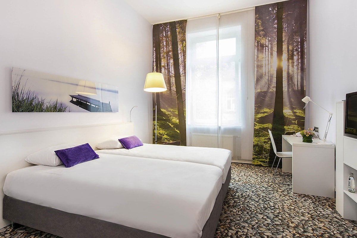 A hotel room with two beds, a desk, a chair, a TV, and a large window with forest-themed curtains.