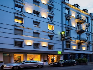 Um hotel com vários andares, janelas iluminadas, entrada visível e placa indicando ser um hotel.