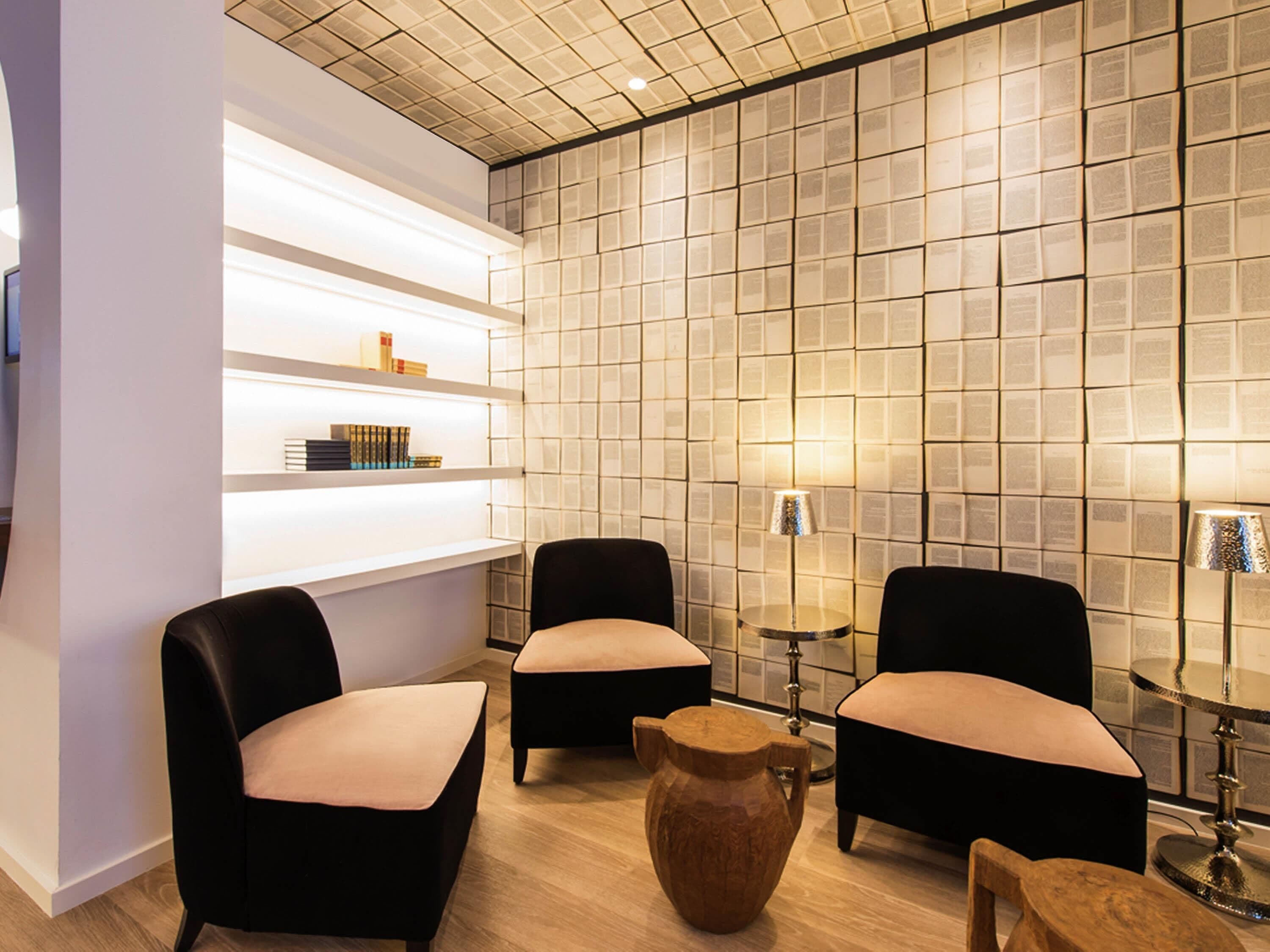 A modern lounge area with a tiled wall, two black armchairs, a small wooden table, and a bookshelf with books.