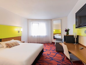 Quarto de hotel com cama, escrivaninha, cadeira, televisão e janela com cortinas.
