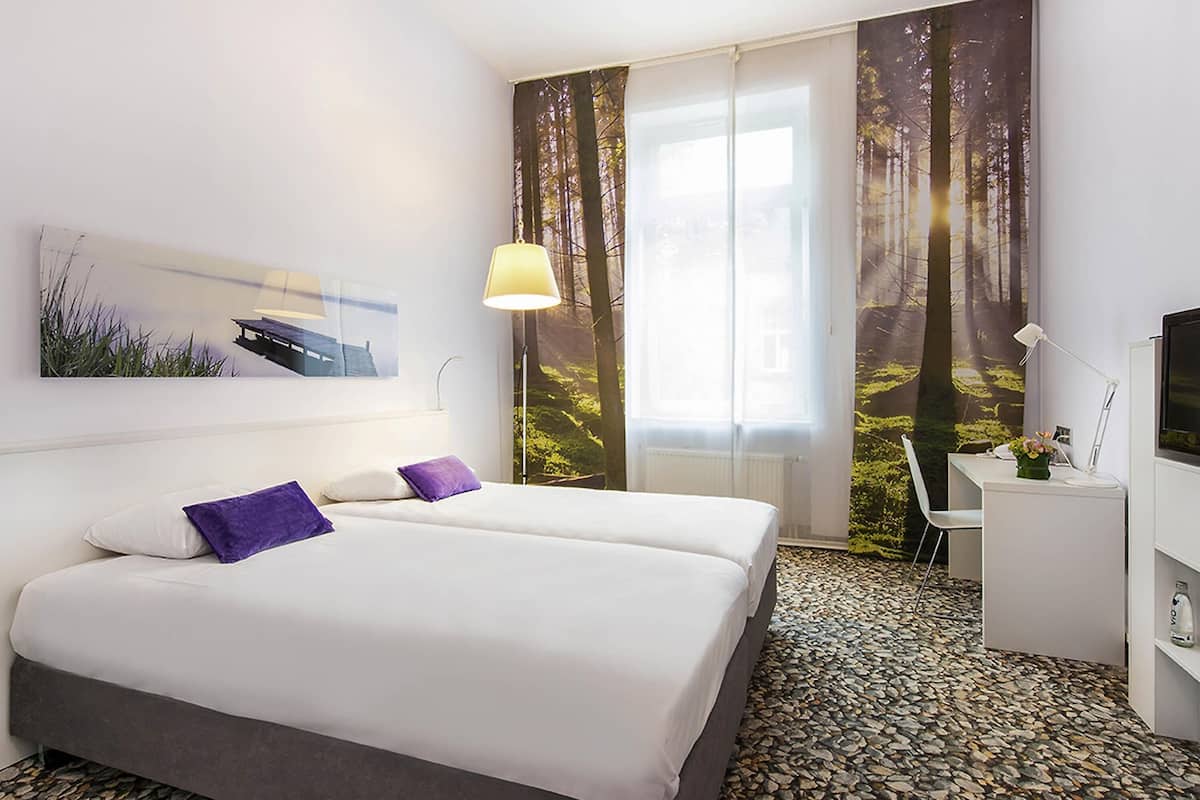 Quarto de hotel com cama, duas almofadas, escrivaninha com cadeira, televisão e janela com cortinas.