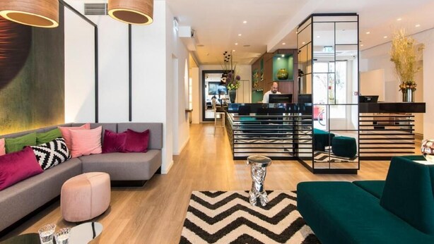 A modern hotel lobby with a grey sofa, a green armchair, and a bar area with a glass partition. - ibis Styles Frankfurt City (Frankfurt)