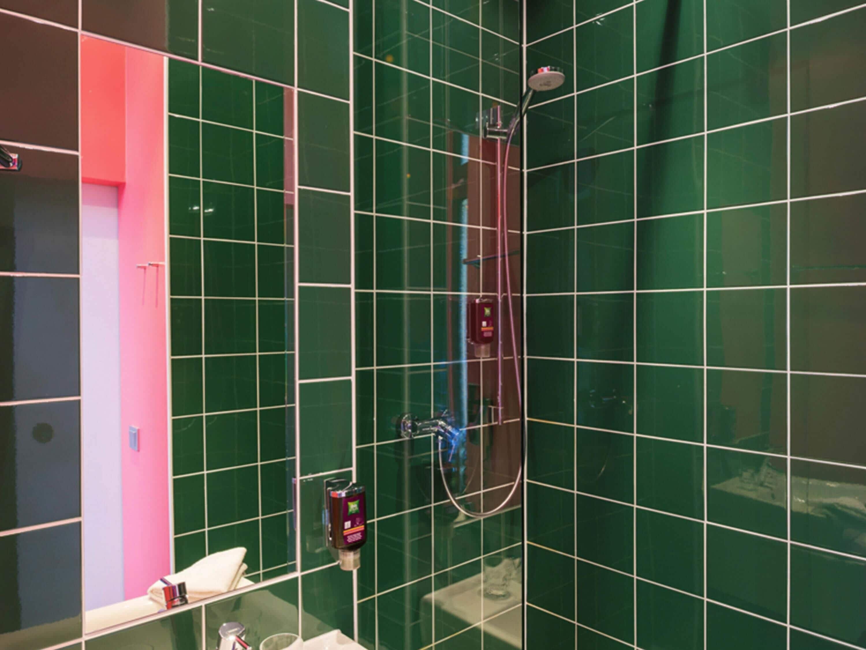 A bathroom with green tiles, a shower, a hand soap dispenser, and a mirror.