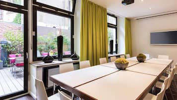 Un salón de conferencias con una larga mesa, sillas blancas, un amplio ventanal con cortinas verdes y un televisor de pantalla plana en la pared.