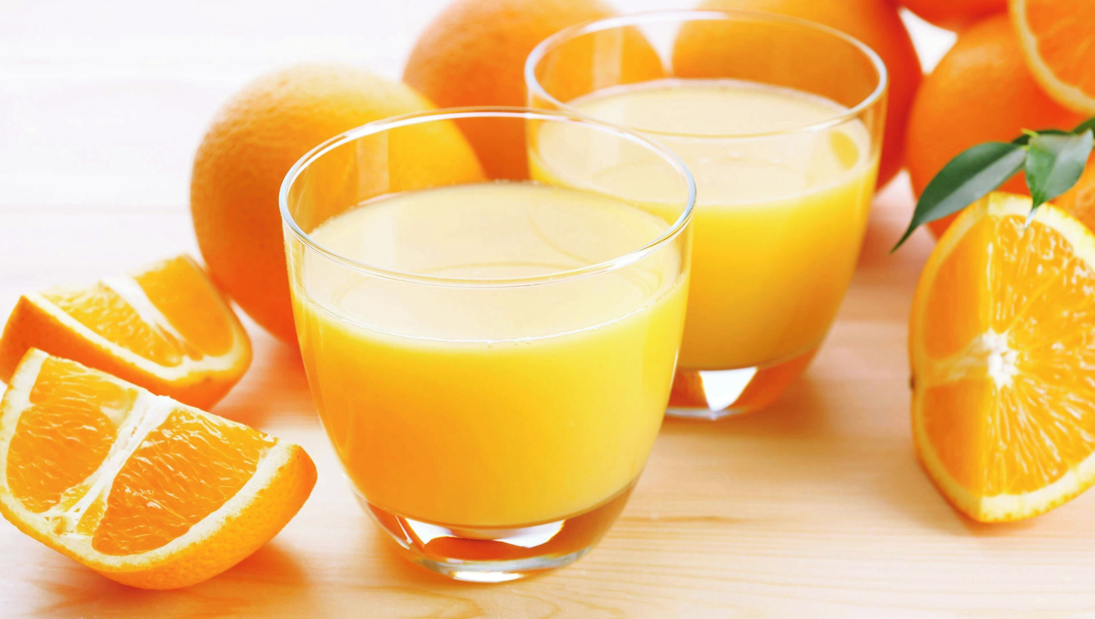 Twee glaasjes sinaasappelsap met hele sinaasappels en schijfjes eromheen.