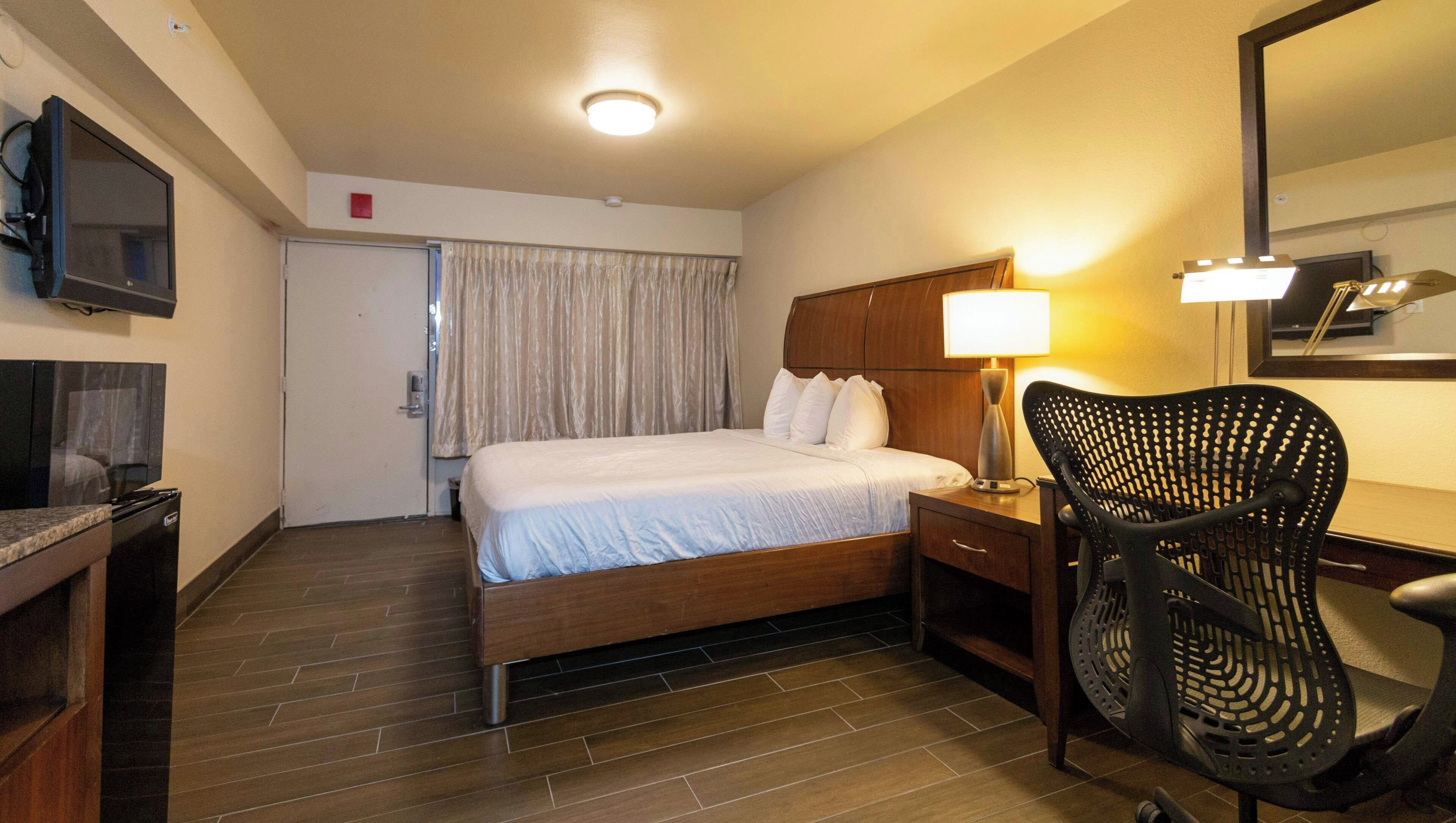 Una habitación de hotel con cama, escritorio con lámpara, silla, televisor y espejo.