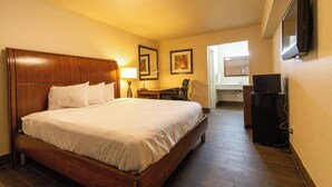 A hotel room with a wooden bed, a desk, a chair, a television, and a bathroom visible through an open door. - Magnuson Extended Stay and Suites Airport Hotel (Irving)