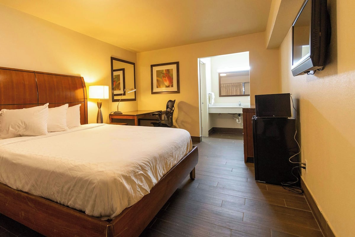 Habitación de hotel con una cama de madera, un escritorio, una silla, un televisor y un baño visible a través de una puerta abierta.