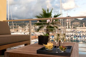 A balcony with a view of a marina, a wooden table with two glasses of champagne, a bowl of olives, and a potted plant.