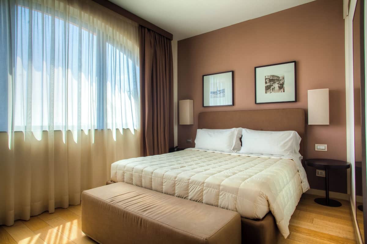 A hotel room with a large bed, a bedside table, a nightstand, a window with sheer curtains, and two framed pictures on the wall.