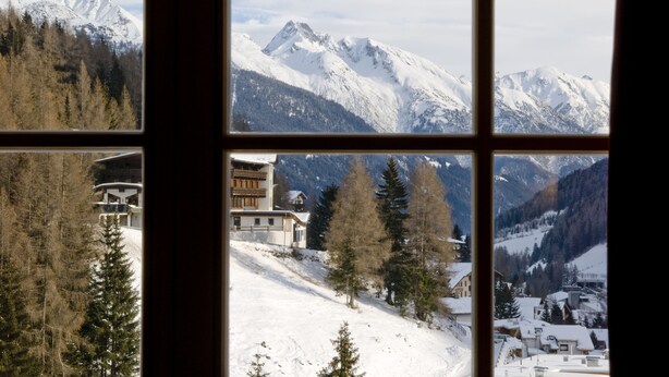 A mountain landscape through a window, including snow-covered peaks, a ski resort, and a building with a wooden facade. - Der Waldhof (Sankt Anton am Arlberg)