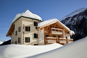 Un edificio cubierto de nieve llamado "Der Waldhof", con balcones de madera y un paisaje montañoso de fondo.