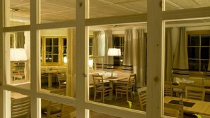 A dining area with wooden tables and chairs, large windows with curtains, and a wooden ceiling. - Der Waldhof (Sankt Anton am Arlberg)