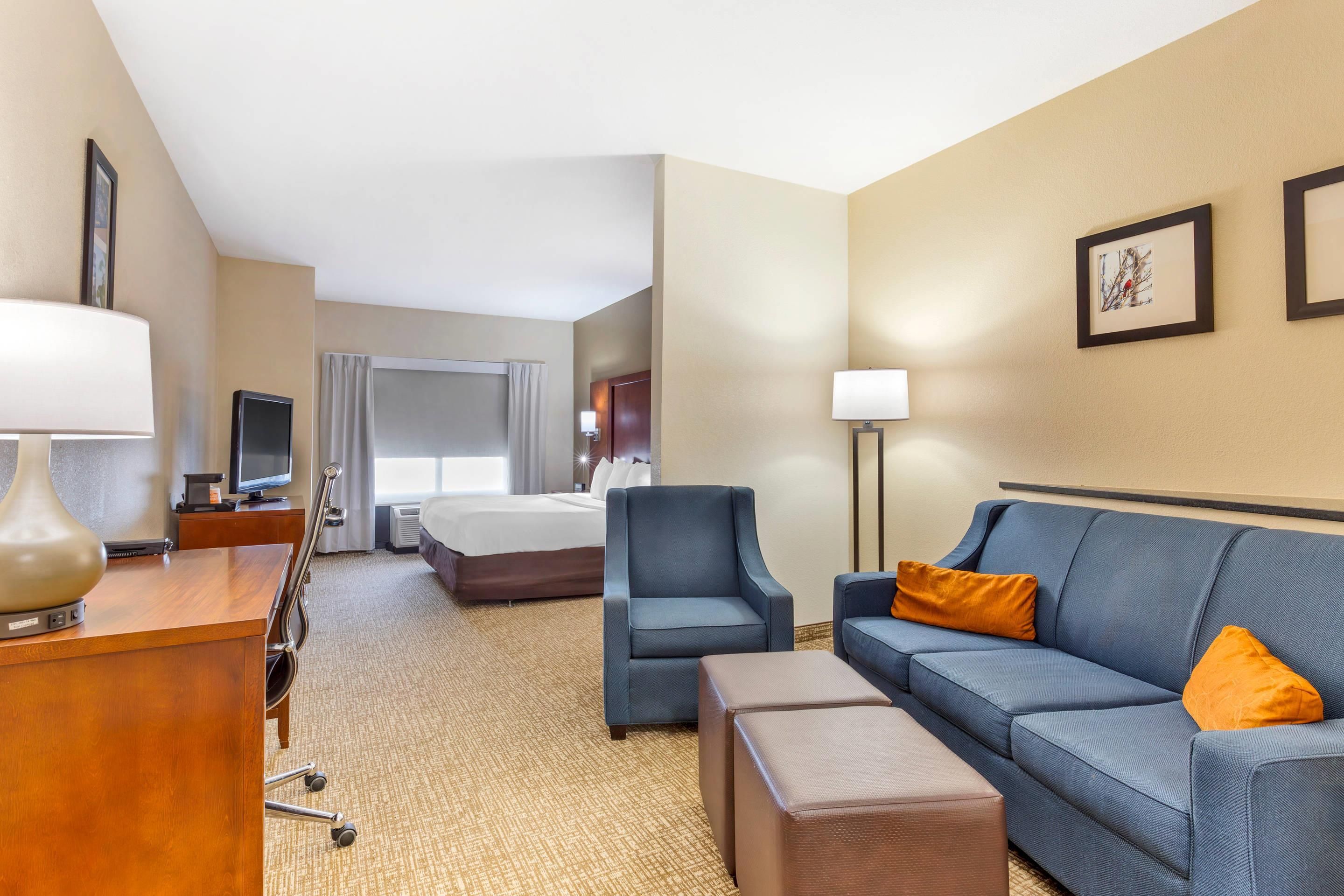 Ein Hotelzimmer mit einem Bett, einem Schreibtisch mit Lampe, einem blauen Sessel, einer Couch mit orangefarbenem Kissen und einem an der Wand befestigten Fernseher.