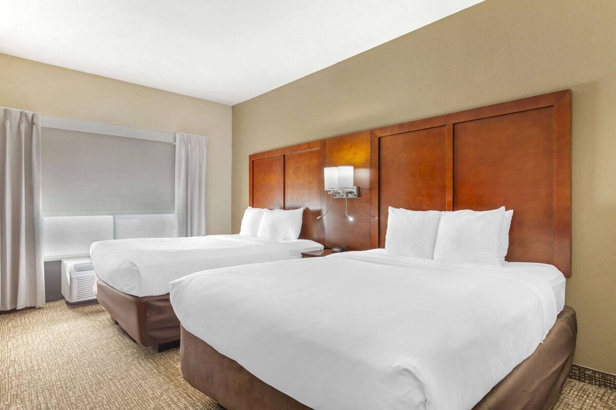 A hotel room with two beds, a large window with curtains, a wall-mounted lamp, and a thermostat.