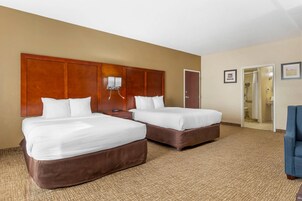 Habitación de hotel con dos camas, cabecero de madera, una mesita de noche, una silla y zona de ducha.