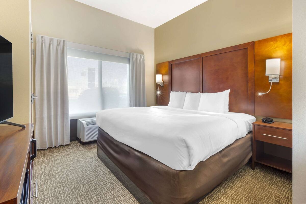 A hotel room with a large bed, a TV, a bedside table, a lamp, and a window with curtains.