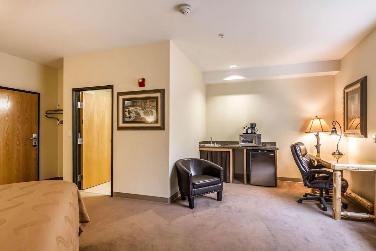 A hotel room with a bed, a desk with a chair, a kitchenette, a framed picture on the wall, and a door.