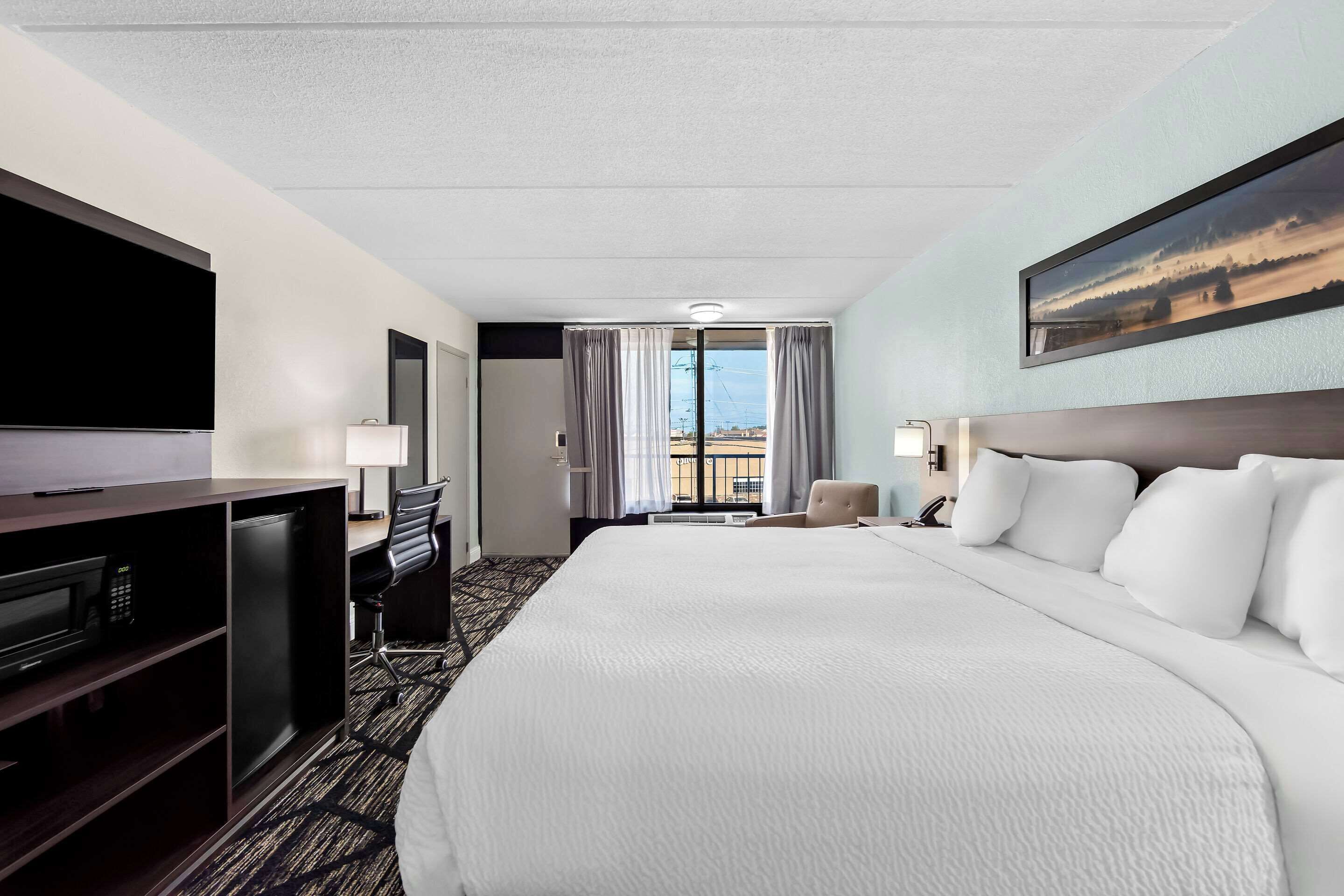 Une chambre d’hôtel dotée d’un grand lit, d’un bureau avec un téléviseur à écran plat, d’une chaise, d’une lampe de bureau et offrant une vue sur l’extérieur grâce à une fenêtre munie de rideaux.