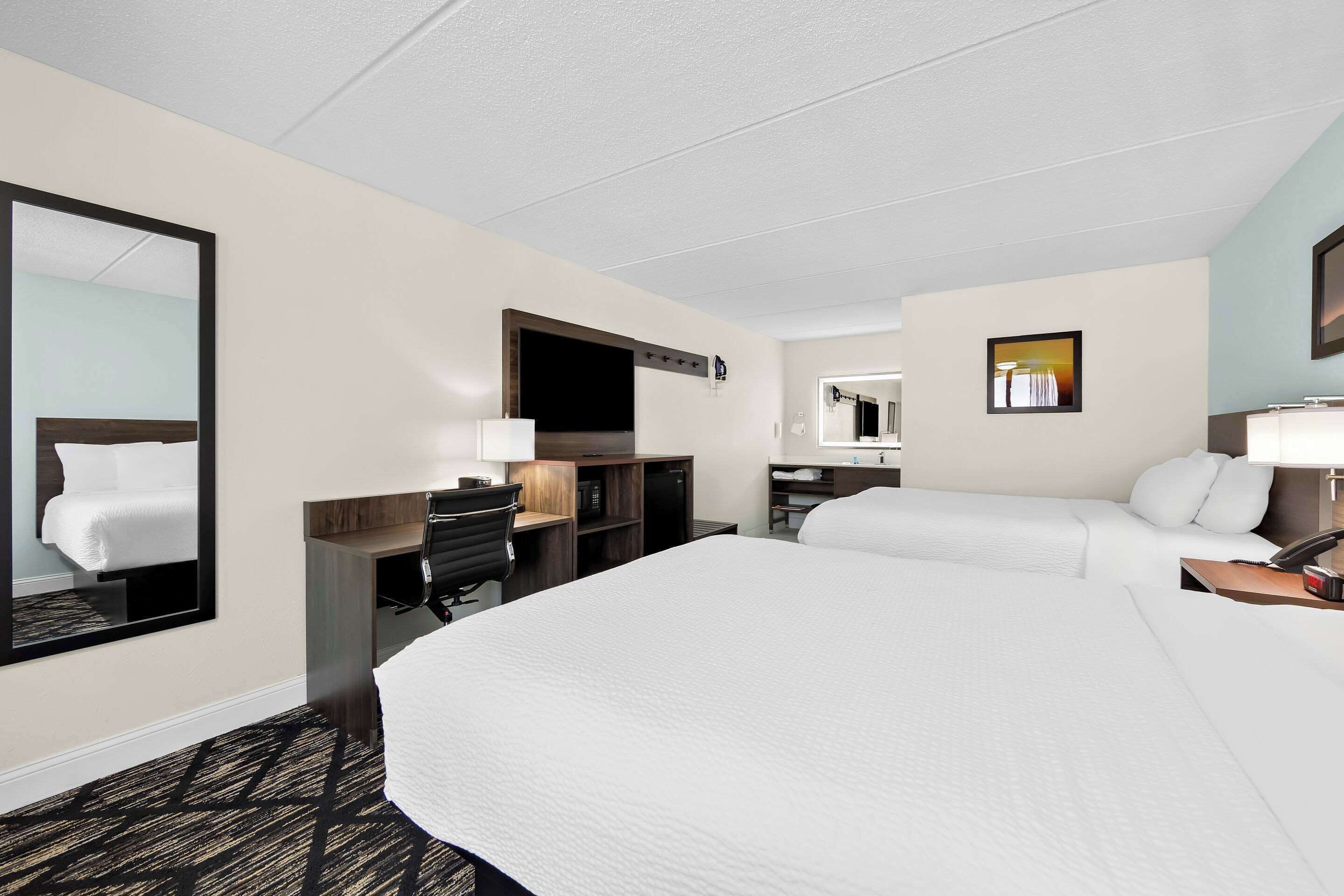 Ein Hotelzimmer mit einem großen Bett, einem Schreibtisch mit Stuhl, einer Schreibtischlampe, einem Flachbildfernseher, einem Spiegel und einem kleinen Tisch mit Telefon und Lampe.